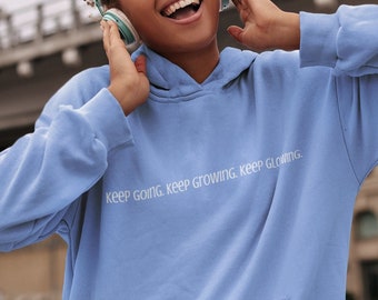 May include: A woman wearing a light blue hoodie with the text "keep going, keep growing, keep glowing" in white. She is wearing headphones and smiling while looking to the right.