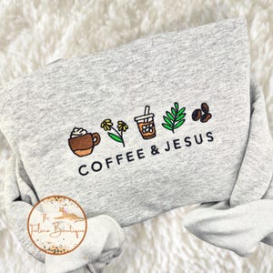 May include: A light gray sweatshirt with embroidered coffee-themed designs and the words "COFFEE & JESUS." The design includes a coffee cup, flowers, iced coffee, a leaf, and coffee beans. The sweatshirt is folded.