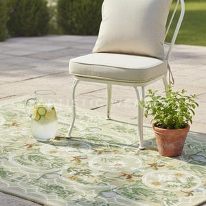 May include: An outdoor scene featuring a white metal chair with a beige cushion and pillow. A green patterned rug, a glass pitcher with lemon and cucumber slices, and a potted plant complete the setting. The scene evokes a relaxing atmosphere.
