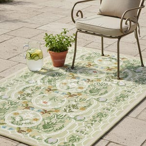 May include: A light green outdoor rug with a floral pattern, featuring a beige border. A metal chair with a beige cushion and pillow sits on the rug. A glass pitcher of water with cucumber slices and a potted plant are also present.