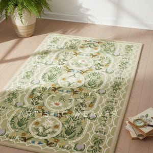 May include: A rectangular, light green rug with a floral pattern. The rug features a repeating design of circular floral medallions and botanical elements. The rug is bordered with a pink trim and is placed on a light wood floor.