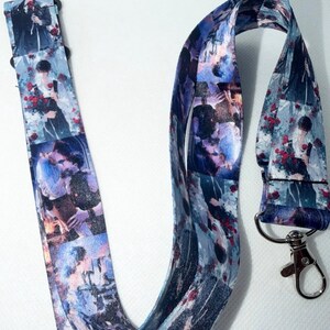 May include: A fabric lanyard featuring a repeating digital print of anime-style characters in romantic poses surrounded by red roses. The strap is predominantly blue and white with a silver-toned metal lobster clasp attached at the bottom end.