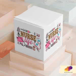 May include: A white cube-shaped notepad with the words "NURSE LIFE" in leopard print, surrounded by medical-themed illustrations. The design includes a stethoscope, syringe, pills, and a prescription bottle. The notepad is on a multi-level surface.