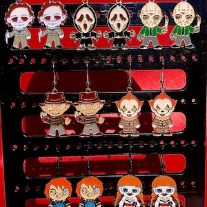 May include: A set of 10 pairs of horror movie character earrings. The earrings are made of metal and feature characters from popular horror movies such as Freddy Krueger, Jason Voorhees, and Pennywise. The earrings are displayed on a black stand.