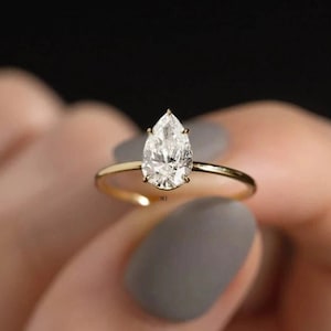 Puede incluir: Un anillo de oro con un diamante en forma de pera. El anillo está en un dedo con esmalte de uñas gris.