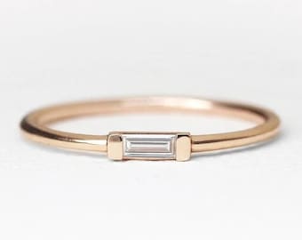 May include: A delicate gold band ring with a single, small, rectangular-shaped diamond set in the center.