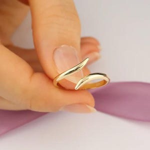 Puede incluir: Un anillo dorado con un diseño abierto único. El anillo presenta dos extremos curvos y puntiagudos que se superponen ligeramente. El anillo se muestra sobre un fondo rosa suave.