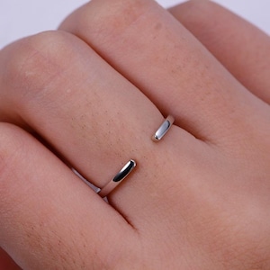 May include: A silver-colored adjustable ring. The ring has a simple, minimalist design with an open band. The ends of the ring are smooth and rounded. The ring is worn on a finger.