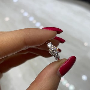 Op de afbeelding: Een zilveren ring met drie ronde diamanten gezet in een kraalbezetting. De ring is op een vinger met rode nagellak.