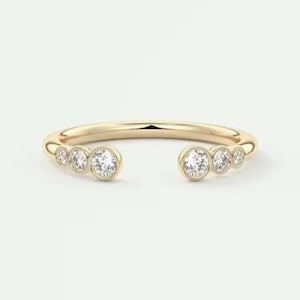 Könnte beinhalten: Ein goldfarbener offener Ring mit sieben runden, klaren Edelsteinen. Die Edelsteine sind in einer Lünettenfassung gefasst, mit drei auf jeder Seite und einem in der Mitte. Der Ring befindet sich auf weißem Hintergrund.