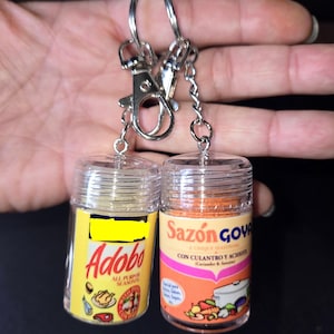 May include: Two miniature spice jar keychains. One jar is yellow with the word "Adobo" and the words "All Purpose Seasoning". The other jar is orange with the words "Sazón Goya" and "Con Culantro y Achiote". Both have silver key rings.