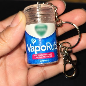 May include: A miniature clear plastic container shaped like a VapoRub jar attached to a silver metal keychain. The label is blue and white with the text VapoRub, COUGH SUPPRESSANT, TOPICAL ANALGESIC, and Ointment printed in white and red.