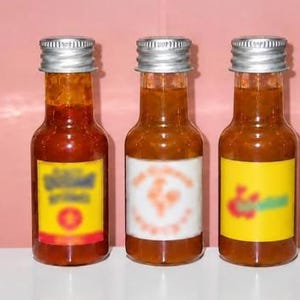 Puede incluir: Tres botellas pequeñas de vidrio llenas de salsa picante de color naranja rojizo, cada una con una tapa de rosca plateada. Las botellas tienen etiquetas rectangulares en amarillo y blanco, que muestran texto y gráficos borrosos.