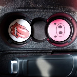 May include: Two round car cup holders with decorative inserts. The left insert is white with a red tongue and lips design. The right insert is pink with a pig face design.