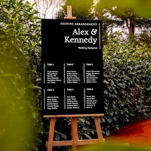 May include: Black chalkboard easel sign with white text for a wedding reception. The sign lists the names "Alex & Kennedy" and the seating arrangement for guests at tables 1 through 6.