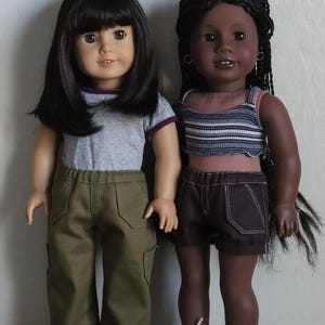 May include: Two dolls wearing casual outfits. The doll on the left has black hair and is wearing a gray t-shirt with a purple trim, green cargo pants, and white sneakers. The doll on the right has dark brown skin, black braids, and is wearing a striped tank top, dark denim shorts, and blue sneakers.