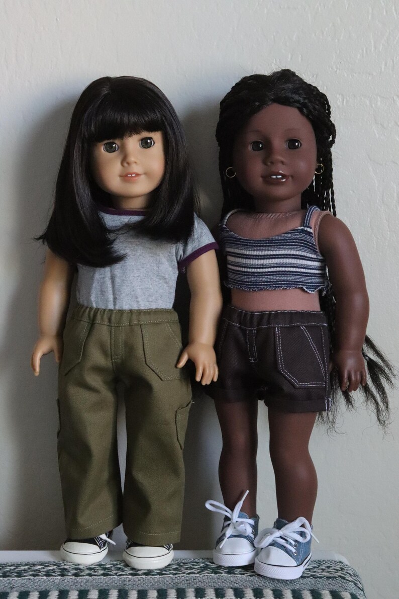 May include: Two dolls wearing casual outfits. The doll on the left has black hair and is wearing a gray t-shirt with a purple trim, green cargo pants, and white sneakers. The doll on the right has dark brown skin, black braids, and is wearing a striped tank top, dark denim shorts, and blue sneakers.