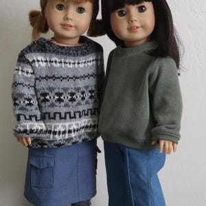 May include: Two dolls are shown, each with a different hairstyle and outfit. One doll wears a gray and white patterned sweater, a denim skirt, gray socks, and navy shoes. The other doll wears a green sweater and blue jeans with white shoes.
