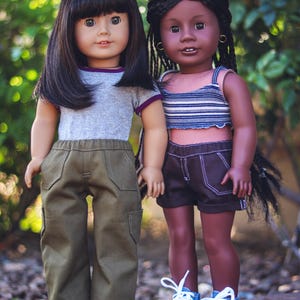 May include: Two dolls, one with black hair and wearing a gray shirt and green pants, and the other with brown hair and wearing a striped shirt and denim shorts. Both dolls are wearing white sneakers.