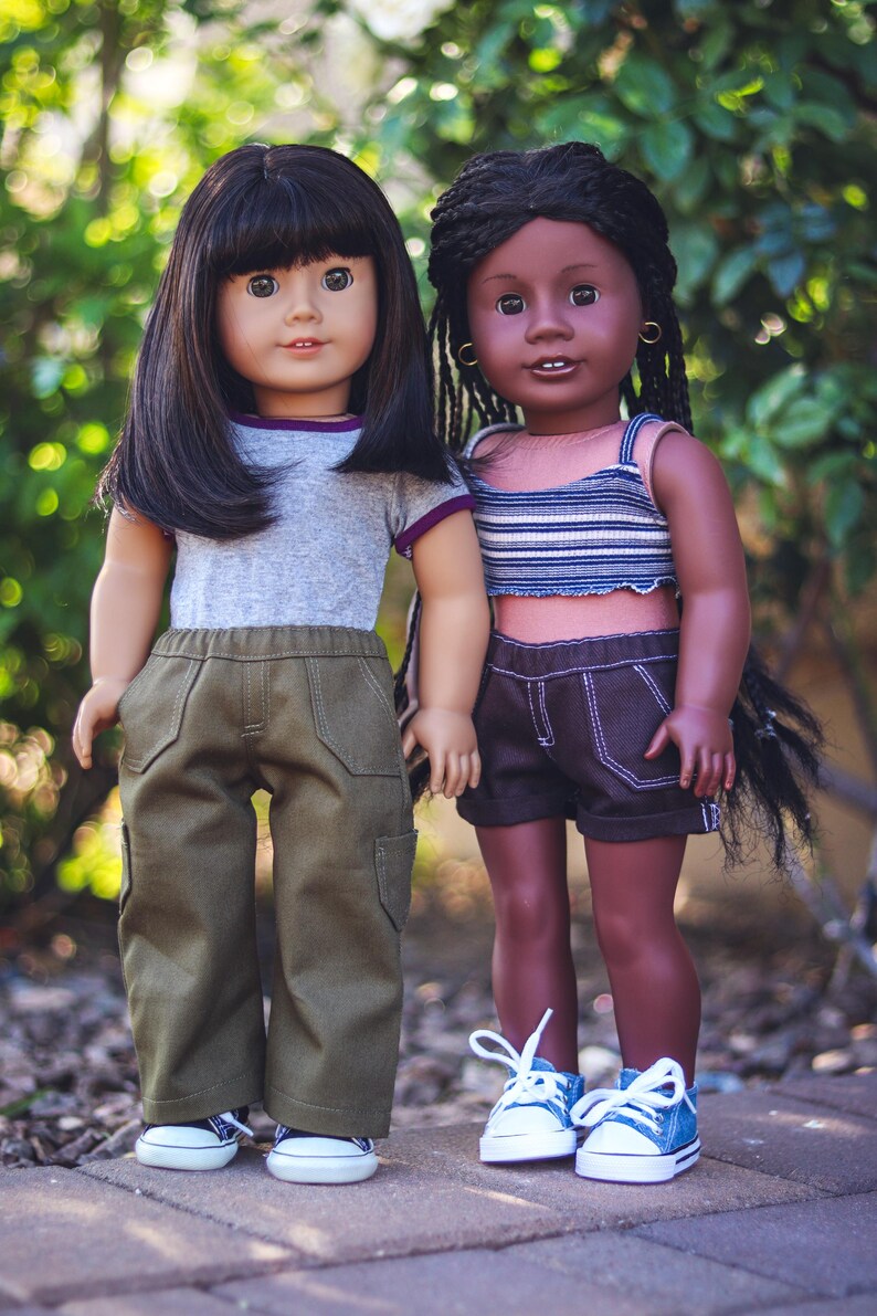 May include: Two dolls, one with black hair and wearing a gray shirt and green pants, and the other with brown hair and wearing a striped shirt and denim shorts. Both dolls are wearing white sneakers.