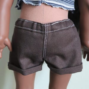 May include: Brown denim shorts with a cuffed hem and white stitching. The shorts are worn by a doll with brown skin.