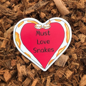Op de afbeelding: Een rood hartvormig bord met een witte rand en een geel slangenontwerp. Het bord zegt "Must Love Snakes".