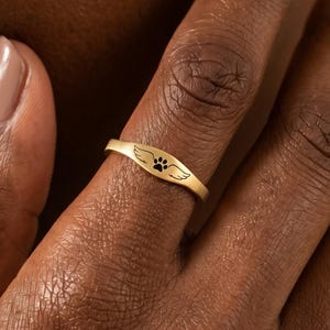 May include: A gold ring featuring a paw print and wing design. The ring is oval and worn on a finger. The paw print is central, flanked by wings. The ring is a piece of jewellery.