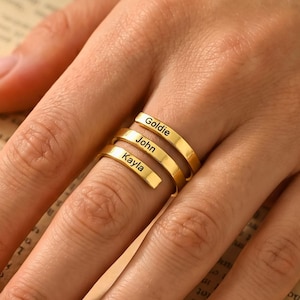 Puede incluir: Un anillo dorado con tres bandas, cada una grabada con un nombre: Goldie, John y Kayla. El anillo se lleva en un dedo, con los nombres claramente visibles. Es una joya personalizada.