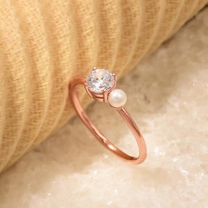 May include: A delicate rose gold ring featuring a single clear stone and a small pearl accent.