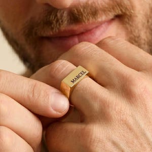 Op de afbeelding: Een goudkleurige zegelring met een plat, rechthoekig bovenoppervlak. De naam MARCEL is in zwarte hoofdletters in het midden van het platte oppervlak gegraveerd. De ring heeft een gladde, gepolijste afwerking en een eenvoudig bandontwerp.