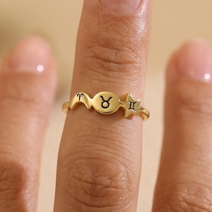 Puede incluir: Un anillo dorado con tres símbolos del zodiaco: Aries, Tauro y Géminis. El anillo se lleva en un dedo, con los símbolos dispuestos en una barra horizontal. El anillo es una pieza de joyería delicada.