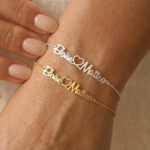 Puede incluir: Dos pulseras delicadas, una plateada y otra dorada, cada una con el nombre "Boise Matteo" en escritura cursiva con un símbolo de corazón. Las pulseras se muestran en una muñeca, mostrando el elegante diseño de la joyería.
