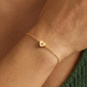 Könnte beinhalten: Ein zartes, goldfarbenes Kettenarmband mit einem kleinen, mittig platzierten herzförmigen Anhänger. Der Anhänger ist mit dem Großbuchstaben A in einer schlichten, klaren Schriftart graviert. Das Armband besteht aus feinen, ineinandergreifenden Gliedern.