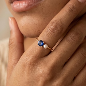 May include: A gold ring featuring a round, deep blue gemstone and a small, white spherical pearl set side-by-side on a thin, polished gold band. The ring is worn on a finger, showcasing the contrast between the vibrant blue stone and the pearl.