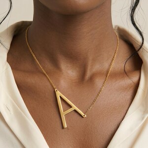 Puede incluir: Un collar dorado con un gran colgante con la letra "A". El colgante está conectado a una delicada cadena. El collar se lleva sobre una camisa de color crema.