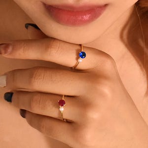 May include: Two gold rings, each featuring a single pearl and a gemstone. The ring on the left has a blue gemstone, while the ring on the right has a red gemstone.