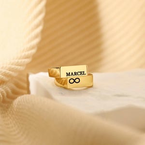 Puede incluir: Dos anillos dorados apilados. El anillo superior tiene grabado el nombre "MARCEL", y el inferior un símbolo de infinito. Los anillos están sobre una superficie blanca, con un fondo dorado suave.