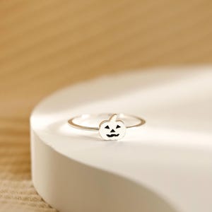 Puede incluir: Un anillo de plata con un pequeño dije de calabaza. La calabaza tiene una cara tallada y un tallo, engarzados en una banda delgada. Un accesorio con temática de Halloween.