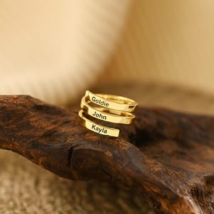 Op de afbeelding: Een goudkleurige spiraalring met de namen "Goldie", "John" en "Kayla" gegraveerd. De ring wordt getoond op een stuk donkerbruin hout. De achtergrond is wazig.