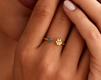 May include: A gold-toned open ring featuring a small blue heart-shaped stone on one end and a gold paw print charm with the letter W engraved on the other end. The ring is designed with a thin, polished band that wraps around the finger.