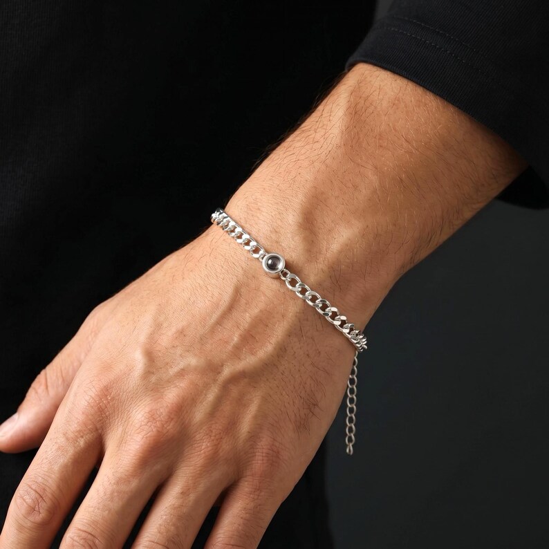 May include: A silver chain bracelet with a small, dark-colored circular accent. The bracelet is made of linked metal pieces and is worn on the wrist. The background is a solid black.