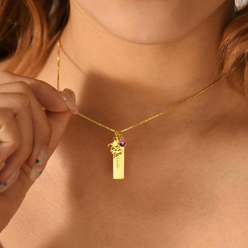 May include: A gold necklace featuring a rectangular pendant engraved with the name "Kardia". A small pink gemstone and a decorative charm are attached to the pendant. The necklace is worn around the neck.
