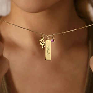 May include: A gold-coloured necklace featuring three charms. One is a vertical rectangle engraved with the name "Charlie". Another is a small, round, purple gemstone. The third charm displays the number "2025".