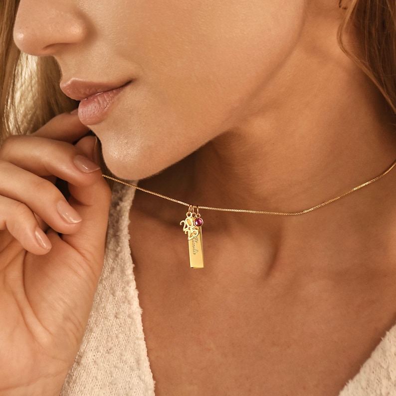 May include: Gold necklace featuring a rectangular pendant with engraved text, a small charm, and a pink gemstone. The necklace is suspended from a fine chain and worn around the neck.