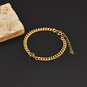 May include: Gold-colored chain bracelet with a small, round, amber-colored stone. The bracelet features a curb chain design and an adjustable clasp. The jewelry is displayed on a black surface with a textured, beige stone in the background.