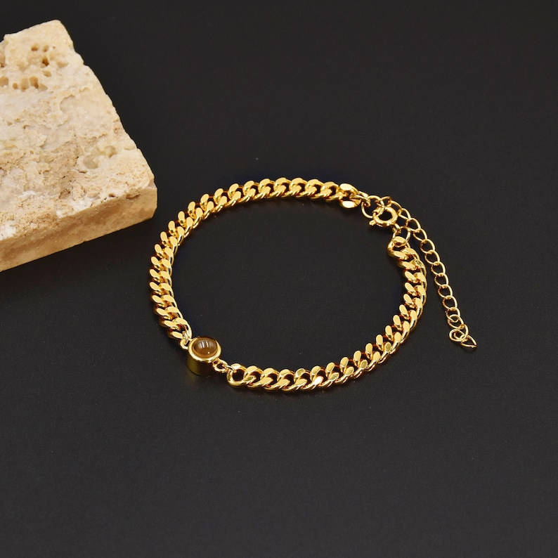 May include: Gold-colored chain bracelet with a small, round, amber-colored stone. The bracelet features a curb chain design and an adjustable clasp. The jewelry is displayed on a black surface with a textured, beige stone in the background.