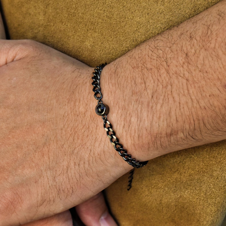 May include: A black chain bracelet with a single, dark-colored gemstone. The bracelet is made of linked metal and is worn on a wrist. The background is a solid, muted brown.