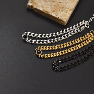 May include: Three chain bracelets in silver, gold, and black, each with a small black stone accent. The bracelets are displayed on a dark surface with a textured beige stone in the background. The chains have a curb link design.