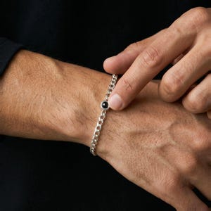 May include: A silver curb chain bracelet with a single black gemstone. The bracelet is on a wrist, with a hand touching the gemstone. The bracelet is a simple, elegant piece of jewelry.