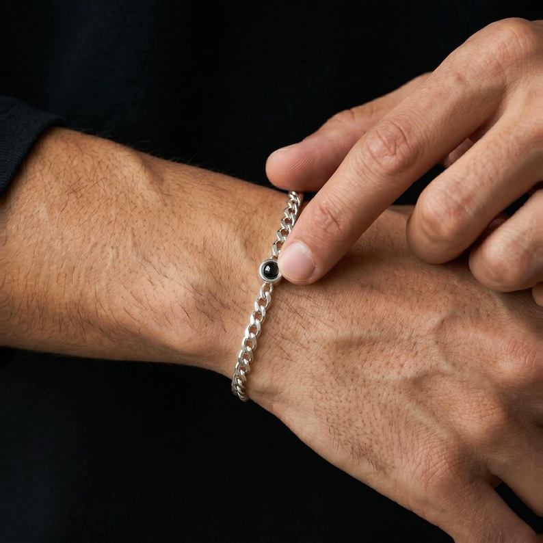 May include: A silver curb chain bracelet with a single black gemstone. The bracelet is on a wrist, with a hand touching the gemstone. The bracelet is a simple, elegant piece of jewelry.